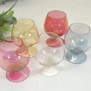 Puede incluir: Cinco copas de vidrio iridiscente en varios colores, incluyendo rosa, amarillo, azul y transparente. Las copas tienen una copa redonda y una base con tallo.