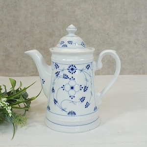 Op de afbeelding: Witte keramische theepot met deksel, versierd met een blauw bloemmotief. De theepot heeft een gebogen tuit en een handvat. Het ontwerp omvat gestileerde bloemen en lijnen. Een klassieker voor het serveren van thee.