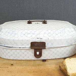 Può includere: Scatola per pane in smalto bianco con un motivo a rete di pollo e una chiusura in metallo arrugginito. La scatola è di forma ovale e ha una maniglia nella parte superiore.