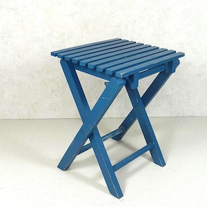 Puede incluir: Un taburete plegable azul con asiento cuadrado de listones y base en forma de X. La estructura de madera tiene un acabado mate con desgaste visible en los bordes. El taburete cuenta con cuatro patas unidas por barras de soporte horizontales.