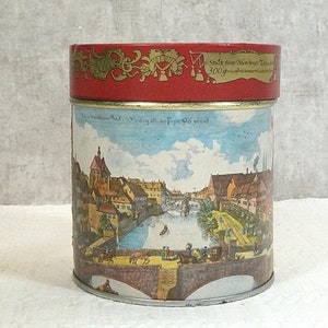 Könnte beinhalten: Eine vintage rote und goldene Dose mit einem Bild einer Stadtlandschaft. Der Text auf der Dose lautet "Stück from Nürnberger Lebkuchen 300g".