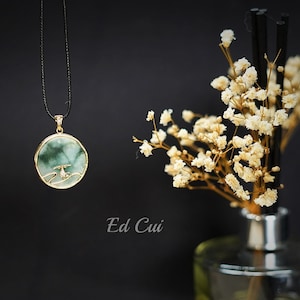 May include: A green jade pendant necklace with a gold setting and a delicate bird design. The necklace is hanging from a black cord and is displayed against a black background.