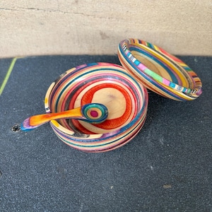 May include: A small, round wooden bowl with a matching lid and a tiny spoon, all crafted from multi-colored, layered wood. The bowl and lid feature concentric rings of vibrant colors including red, blue, yellow, and green, creating a striped pattern.