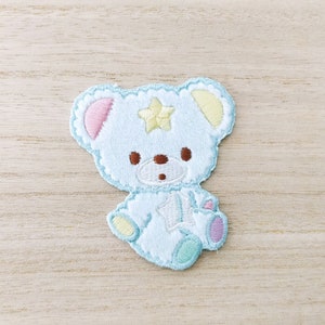 May include: A light blue embroidered plush teddy bear with a yellow star on its chest. The bear has pink and yellow ears and a brown nose.