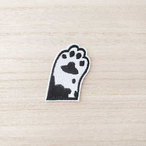 May include: A black and white embroidered cat paw patch. The patch shows a detailed paw print design with visible pads and claws. The patch is set against a light wood grain background, accentuating the contrasting colours.