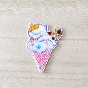May include: An embroidered iron-on patch featuring a white cat with orange and brown markings sitting on top of a pink and purple ice cream cone with white, blue, and pink stars.
