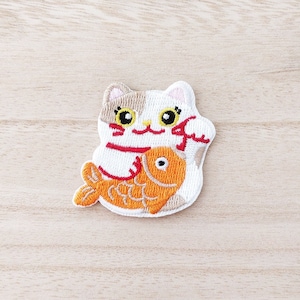 Peut inclure: Écusson brodé représentant un chat Maneki-neko tenant un poisson rouge. Le chat a un corps blanc avec des marques marron et beige, un ruban rouge et un visage souriant. Le poisson rouge est orange. L'écusson est sur un fond de grain de bois clair.