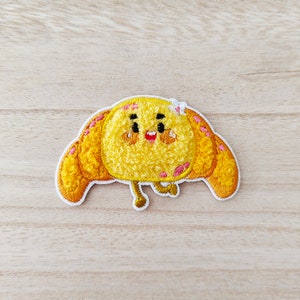 May include: A yellow embroidered patch of a cartoon croissant with a smiling face. The croissant has pink and white details and a white flower on top.
