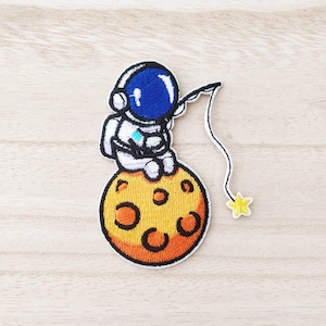 May include: An embroidered patch depicting an astronaut fishing from the moon. The astronaut is wearing a white spacesuit with a blue helmet. The moon is yellow with orange craters. A small yellow star is at the end of the fishing line.