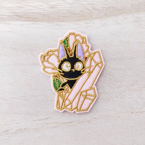 Peut inclure: Chat noir aux grands yeux qui regarde depuis une pochette en forme de cristal rose et or avec des accents floraux.