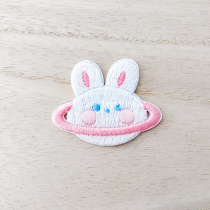 May include: White embroidered iron-on patch of a bunny rabbit with pink ears and a pink ring around it, resembling a planet.