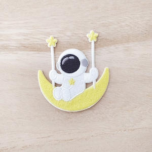 May include: Embroidered patch showing a white astronaut seated on a yellow crescent moon. The astronaut holds two star-shaped wands. The patch has a whimsical, space-themed design.