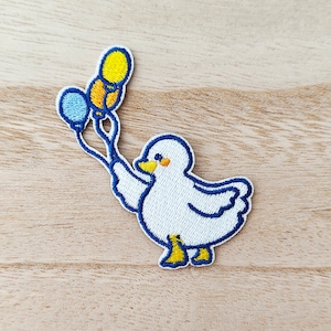 Peut inclure: Patch brodé blanc représentant un canard tenant trois ballons. Les ballons sont bleus, jaunes et orange. Le canard a un bec et des pieds jaunes.