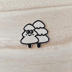 May include: An embroidered patch featuring two stylised evergreen trees in a light cream colour with thick black outlines. The left tree has a simple face with two oval eyes and a small curved mouth, while both trees have a layered, rounded shape.