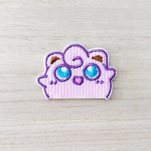 May include: A pink and purple embroidered patch of a cartoon character with blue eyes and a pink heart. The character has a curly brown swirl on its head.