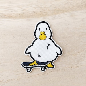 May include: White embroidered patch of a duck wearing a yellow beak and feet, riding a black skateboard with yellow wheels.