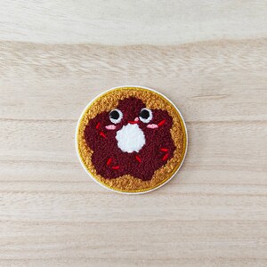 May include: A brown and white embroidered donut with red sprinkles and a happy face. The donut is on a light brown wooden background.