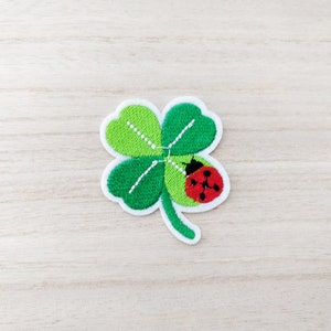 May include: A green four-leaf clover with a red ladybug on one of the leaves. The clover is embroidered on a white fabric.