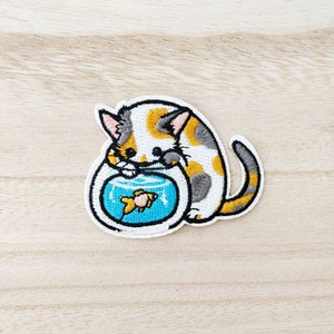 May include: A calico cat with orange, gray, and white markings is looking into a fishbowl. The fishbowl is blue and has a gold fish inside. The cat is embroidered on a white fabric.
