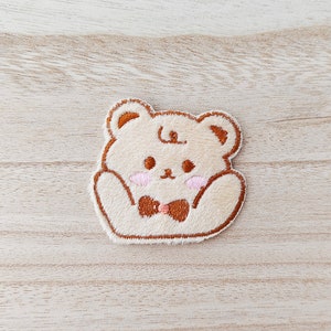 May include: A brown and beige embroidered plush bear patch with a brown bow tie. The bear has a cute expression with pink cheeks.