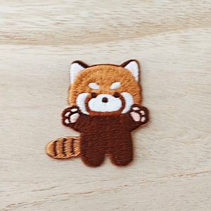 May include: Embroidered patch of a cartoon red panda, with a brown body, white face, and fluffy tail. The patch has detailed stitching and is set against a light wooden surface.
