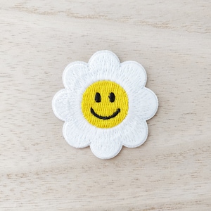 May include: White embroidered flower patch with a yellow smiley face in the centre.