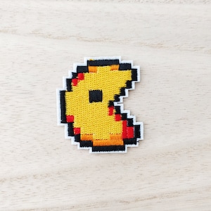 May include: A yellow, pixelated Pac-Man patch with a black outline and a white border. The patch features red accents and a black square in the centre, reminiscent of the classic arcade game character.