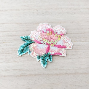 Peut inclure: Une fleur brodée rose et blanche avec des feuilles vertes et des paillettes irisées. La fleur est une rose avec un design détaillé.