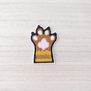 May include: Embroidered brown paw print with pink paw pads and yellow and brown stripes. The paw print is outlined in black.