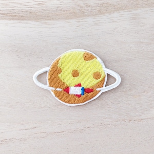 May include: An embroidered patch depicting a yellow and orange planet with a white ring around it. A small red and blue rocket is on the planet.