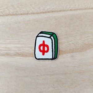 Peut inclure: Un écusson brodé rectangulaire représentant une tuile de mah-jong blanche avec une bordure verte et un caractère chinois rouge au centre. L'écusson présente un contour noir et une surface texturée créée par des points de broderie denses.
