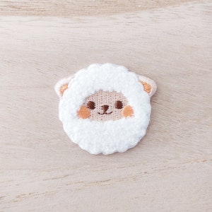 May include: Embroidered patch featuring a cute sheep design. The sheep has a white fluffy face, light brown ears, and rosy cheeks. The patch is a whimsical accessory for clothing or bags.