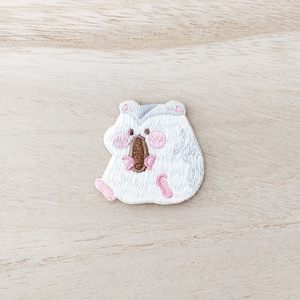 May include: White embroidered iron-on patch of a hamster holding a brown nut in its paws. The hamster has pink cheeks and gray ears.