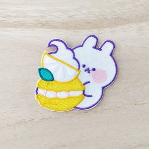 May include: An embroidered white bunny holding a yellow lemon with a green leaf. The bunny has purple outlines and pink cheeks.