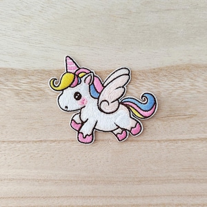 Peut inclure: Écusson brodé d'une licorne blanche avec une corne, des ailes et des sabots roses. La licorne a une crinière jaune et une queue arc-en-ciel. Le patch est bordé de noir et posé sur un fond clair en bois.