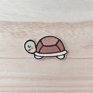 May include: Embroidered turtle patch with a light green head, a brown shell, and a white and light green border. The turtle has a smiling face with a small orange blush. Ideal for adding to clothes or accessories.