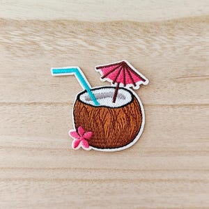 May include: Embroidered patch of a coconut drink with a blue straw, pink umbrella, and a pink flower. The coconut is brown with a white interior. The patch has a white border.