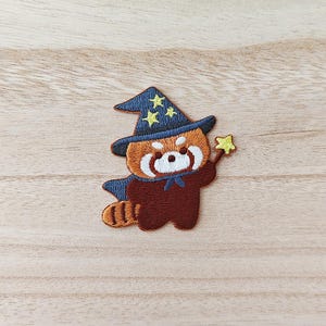 May include: Embroidered patch of a red panda in a wizard costume. The patch shows a red panda wearing a blue wizard hat with yellow stars, a blue cape, and holding a yellow star wand. The panda is brown and white.