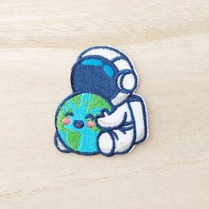 Peut inclure: Écusson brodé représentant un astronaute de dessin animé en combinaison spatiale blanche embrassant une Terre souriante. Le casque de l'astronaute est bleu, et la Terre a des continents verts et des océans bleus. L'écusson est sur une surface en bois clair.