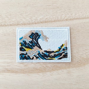 May include: White embroidered patch with a blue, black, and yellow wave design. The wave is a stylized depiction of the Great Wave off Kanagawa, a famous Japanese woodblock print.