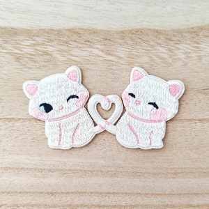Peut inclure: Écusson brodé représentant deux chats blancs de dessin animé avec des détails roses, leurs queues formant un cœur. Les chats sourient, créant un design mignon et romantique, idéal pour les vêtements.