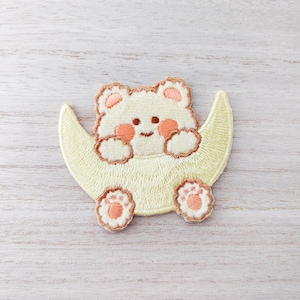May include: A yellow embroidered patch featuring a white bear sitting inside a crescent moon. The bear has brown eyes and a small pink blush on its cheeks.
