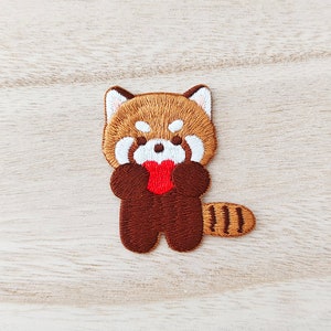 May include: Embroidered patch of a red panda holding a red heart in its paws. The panda is brown with white facial markings and a striped tail.