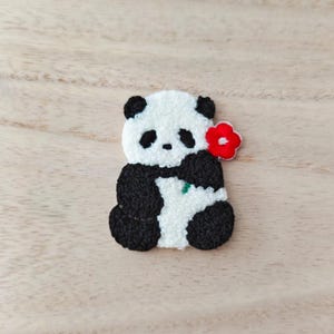 Peut inclure: Écusson fait main représentant un panda noir et blanc tenant une fleur rouge. Le patch est réalisé avec des détails brodés et présente une texture. Le fond est en bois clair, mettant en valeur le motif.