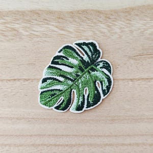 May include: Embroidered patch with a green and white Monstera leaf design. The patch has a white border and is set against a light wood grain background. The leaf's details are intricately stitched, showcasing a tropical plant theme.