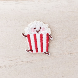 May include: Embroidered patch featuring a smiling popcorn character with a red and white striped container. The popcorn has a fluffy white top, pink cheeks, and brown arms and legs. This patch is perfect for adding a fun touch to clothing or accessories.