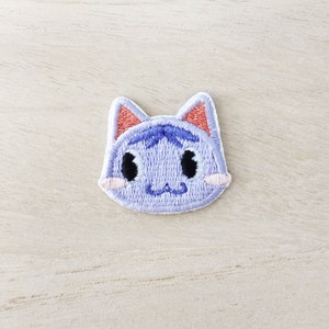 May include: An embroidered patch featuring a cat with a surprised expression. The cat is purple and pink, with large black eyes, a pink nose, and a small pink mouth.