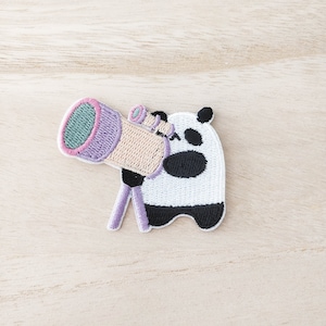 May include: An embroidered iron-on patch featuring a panda bear looking through a telescope. The panda is white with black ears, eyes, and feet. The telescope is purple, pink, and beige.