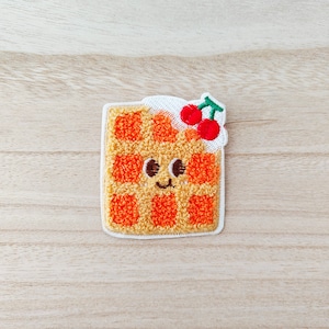May include: An embroidered patch shaped like a waffle with a smiling face and two red cherries on top. The waffle is orange and yellow, and the cherries have green stalks.