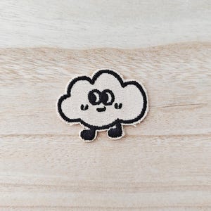 Peut inclure: Un écusson beige en forme de nuage avec un contour noir et un visage souriant. L'écusson a deux yeux noirs, une petite bouche noire et deux petits pieds noirs. L'écusson est sur une surface en bois clair.